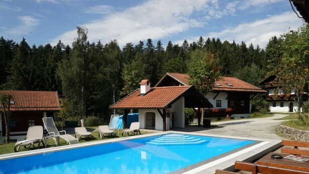 Una piscina con sillas de descanso y un banco de madera, rodeada de casas tradicionales con tejados de tejas rojas, frente a un bosque.