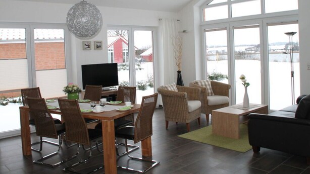 Ein geräumiges Wohnzimmer mit Essbereich, großen Fenstern, einem Flachbildfernseher, Korbmöbeln und Blick auf eine verschneite Landschaft.