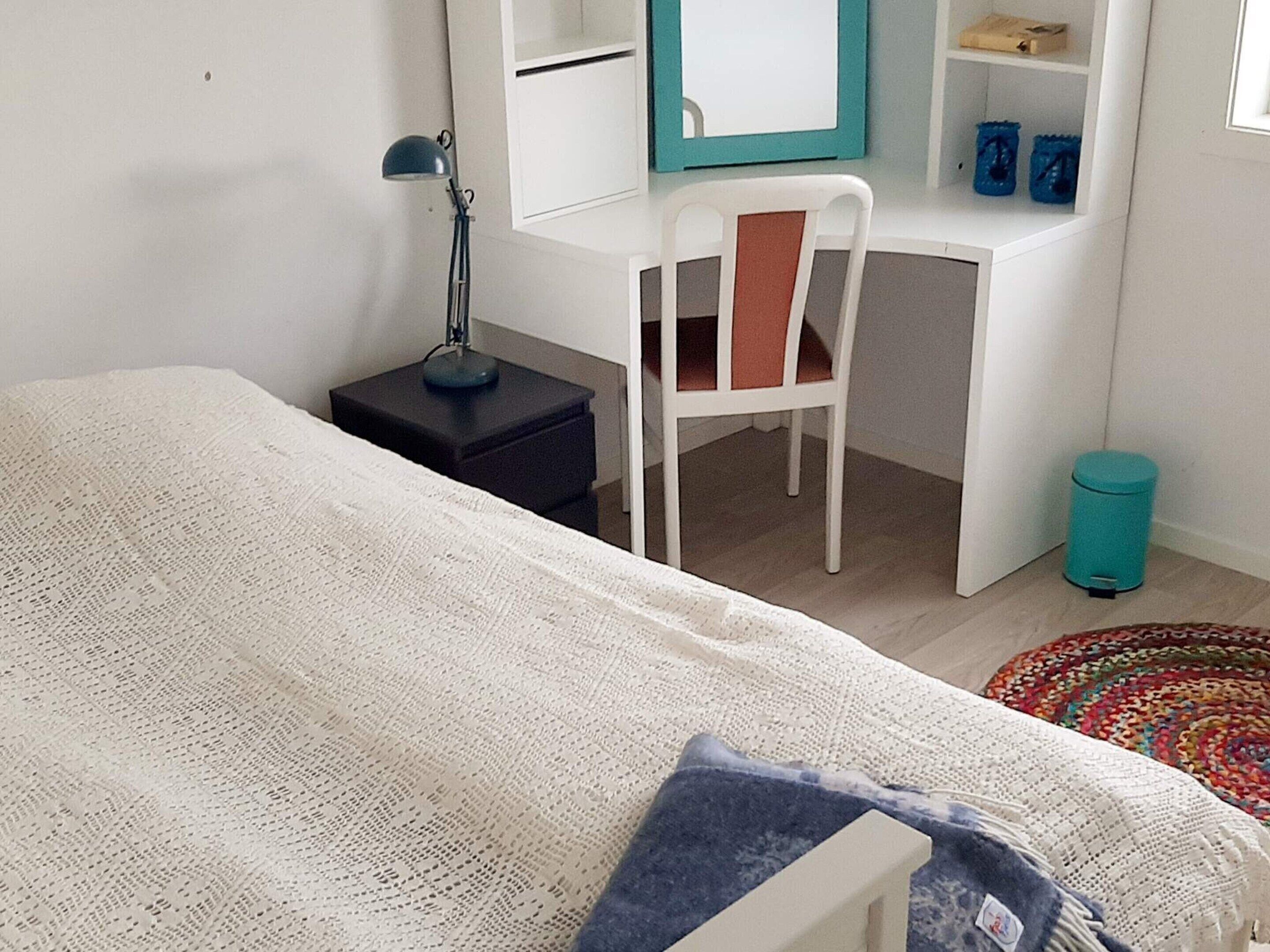 Een slaapkamer met een bed, een bureau, een stoel, een lamp en een prullenbak.