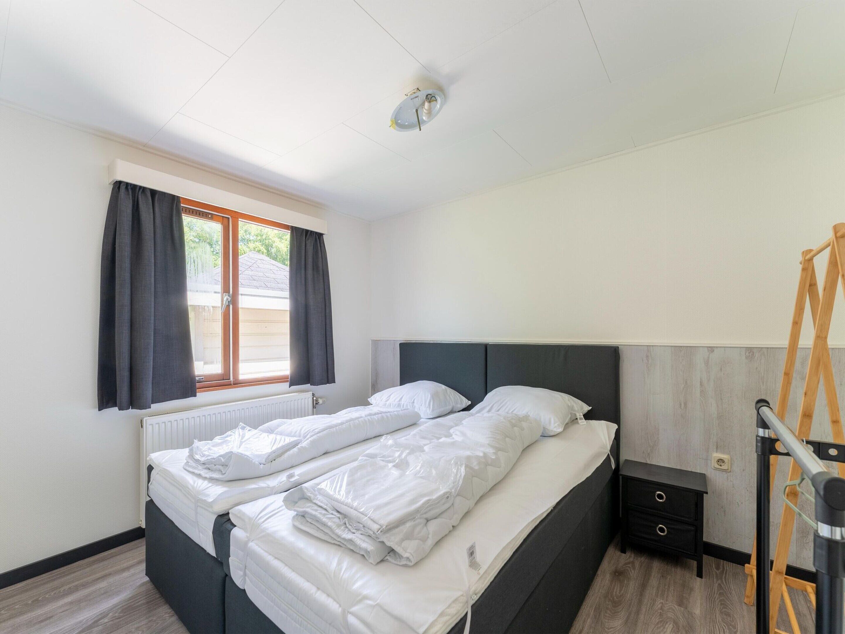 A double bed with white bedding, a window with dark curtains, a radiator, a small bedside table with drawers, and a wooden ladder.