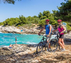 A rocky beach with clear turquoise water, people swimming and sunbathing, and two cyclists with bicycles. - Cosy Apartment in Istria (Vodnjan)