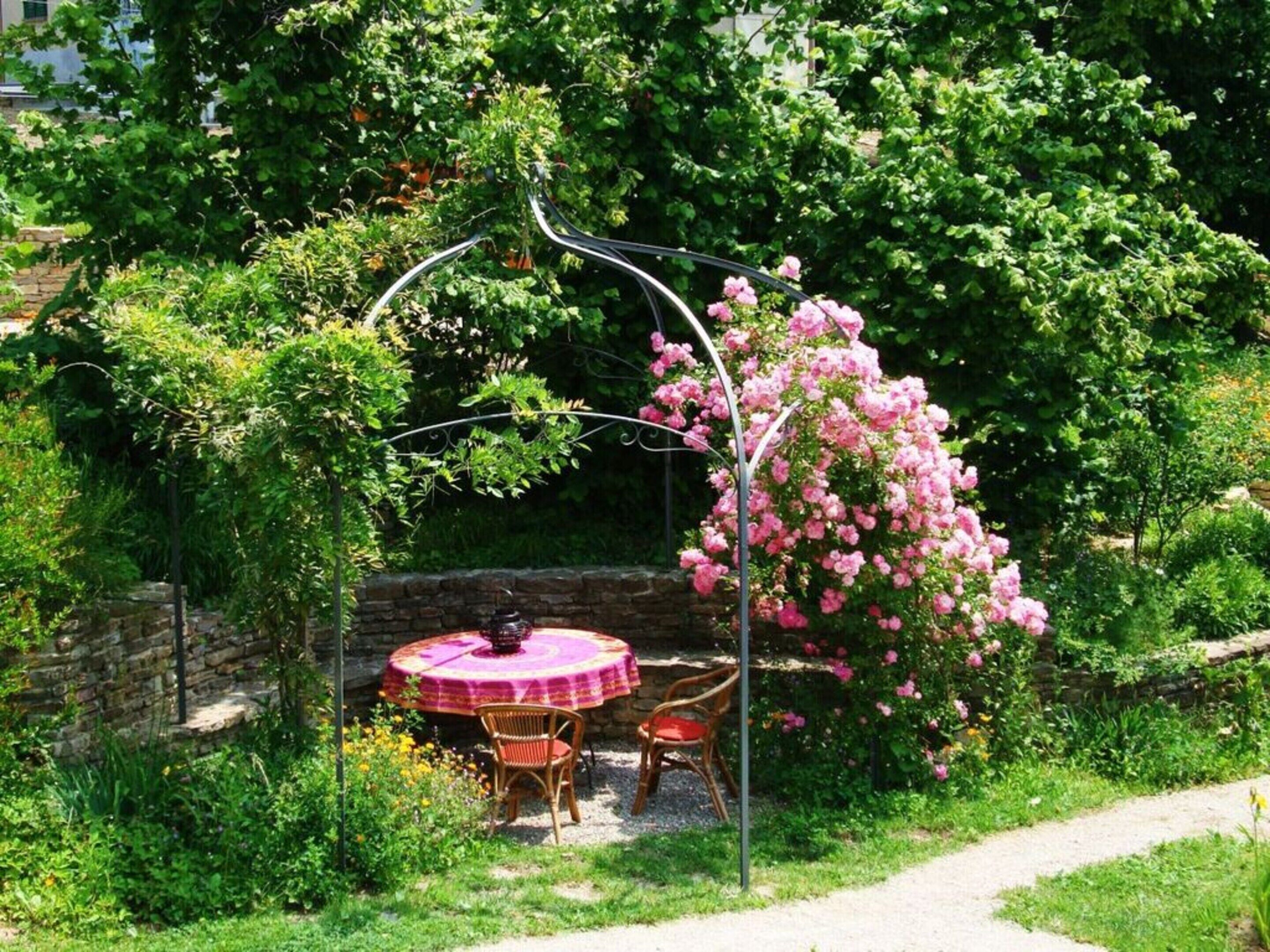 A garden patio with a round table and chairs, surrounded by lush greenery and flowering plants.