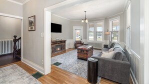 A spacious living room with a fireplace, a large sofa, a vintage trunk, and a view of the outside through large windows. - Gettysburg Home sleeps 10 with parking and Game Room.  Just off the square. (Gettysburg)