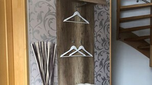 Een houten opbergkast met planken en een kledingroede, een witte kussens en een decoratieve vaas met takken.