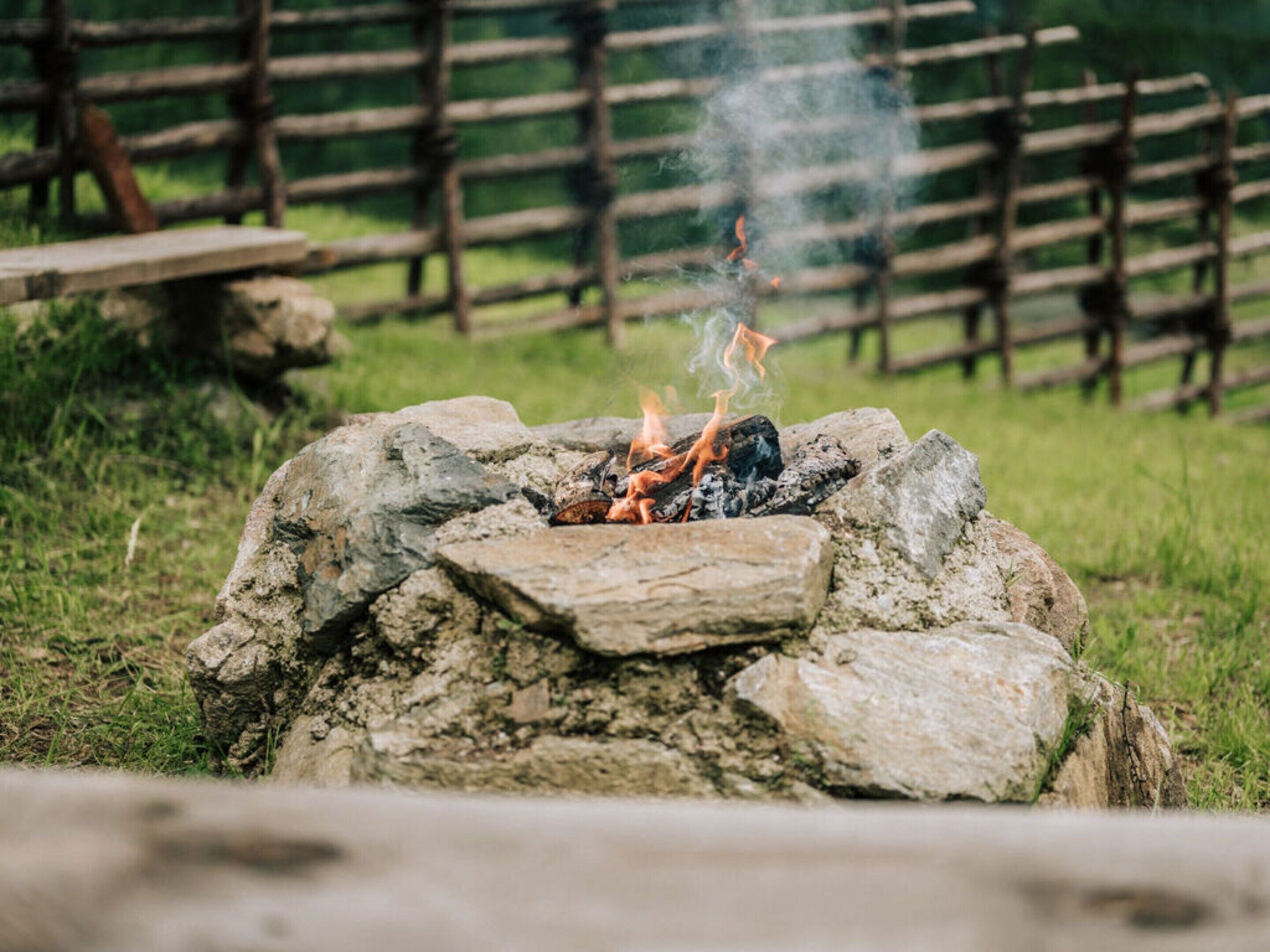 Una fogata con rocas y leña, rodeada por una cerca de madera y un área cubierta de hierba.