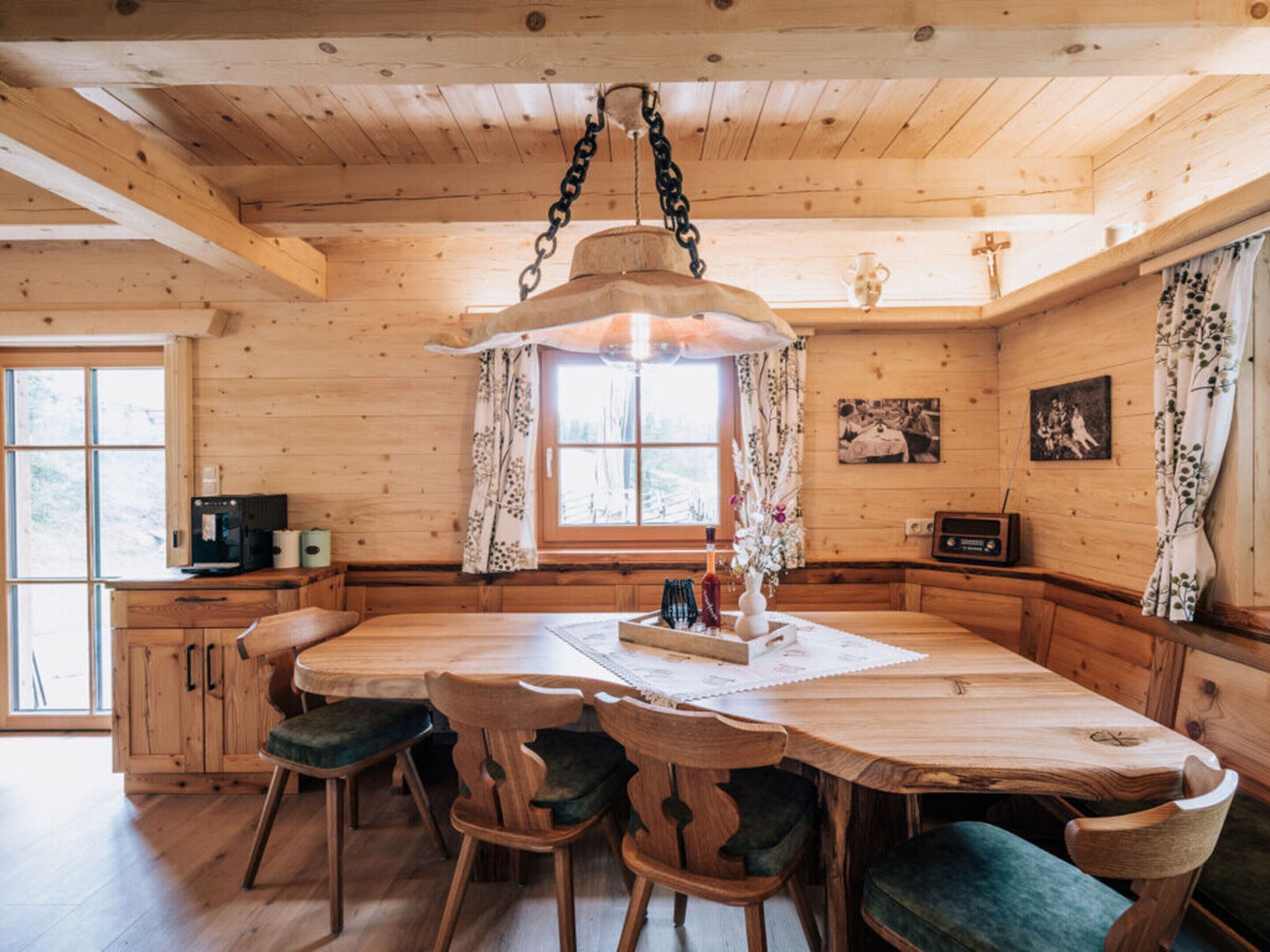Interior de una cabaña de madera con una gran mesa de comedor, sillas, una lámpara colgante, una ventana con cortinas y un radio sobre un mueble de madera.