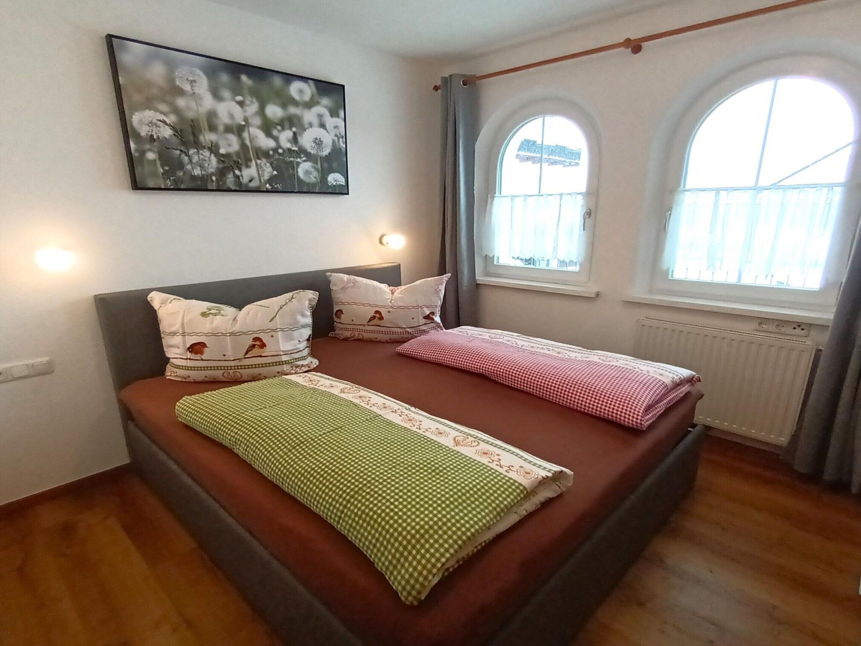 Une chambre avec un lit simple, deux oreillers, une couverture verte et un tableau représentant des fleurs accroché au mur.
