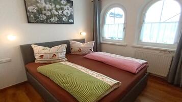 A bedroom with a single bed, two pillows, a green blanket, and a painting of flowers on the wall.
