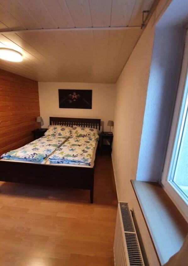 A bedroom with a wooden floor, a bed with floral bedding, a nightstand, a wall-mounted lamp, and a window with a view.