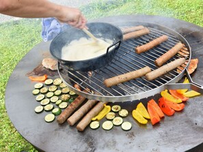 A round outdoor grill with sausages, zucchini slices, and bell peppers being cooked.