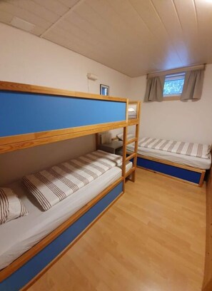 A dormitory room with two bunk beds, wooden flooring, a small window with curtains, and a wall-mounted light fixture.
