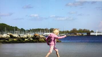 Children can play on the pebbled beach, with calm waters and boats docked in the background.