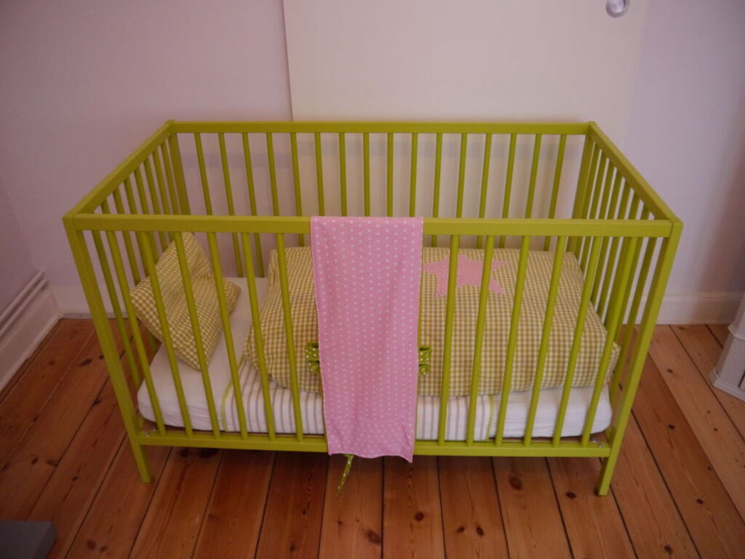 A green baby crib with a pink cloth, checkered bedding, and wooden flooring.
