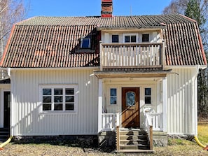 Uma casa de dois andares com telhado de telhas vermelhas, revestimento branco e varanda frontal com porta de madeira e degraus.