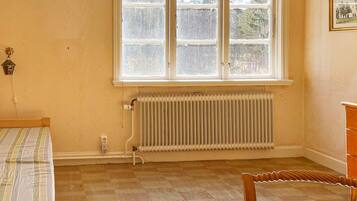 Quarto com radiador, janela vista para árvores, cama e cadeira de madeira.