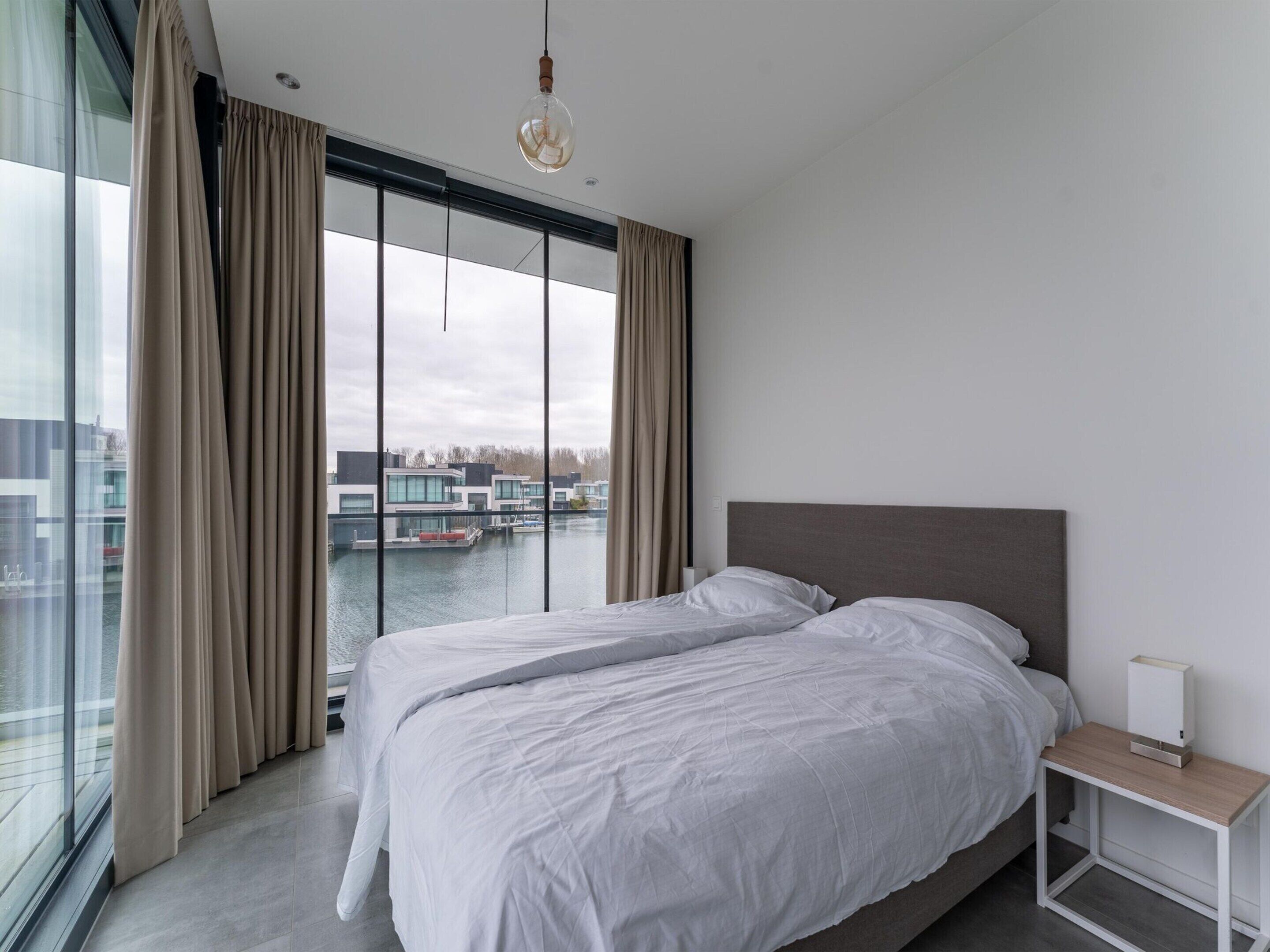 Een moderne slaapkamer met een groot bed, een nachtkastje en uitzicht op een waterpartij en gebouwen.
