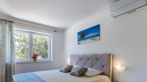 A bedroom with a bed, a nightstand, a window with curtains, a wall-mounted air conditioner, and a painting of a beach scene. - Large Holiday Home With Swimming Pool (Labin)