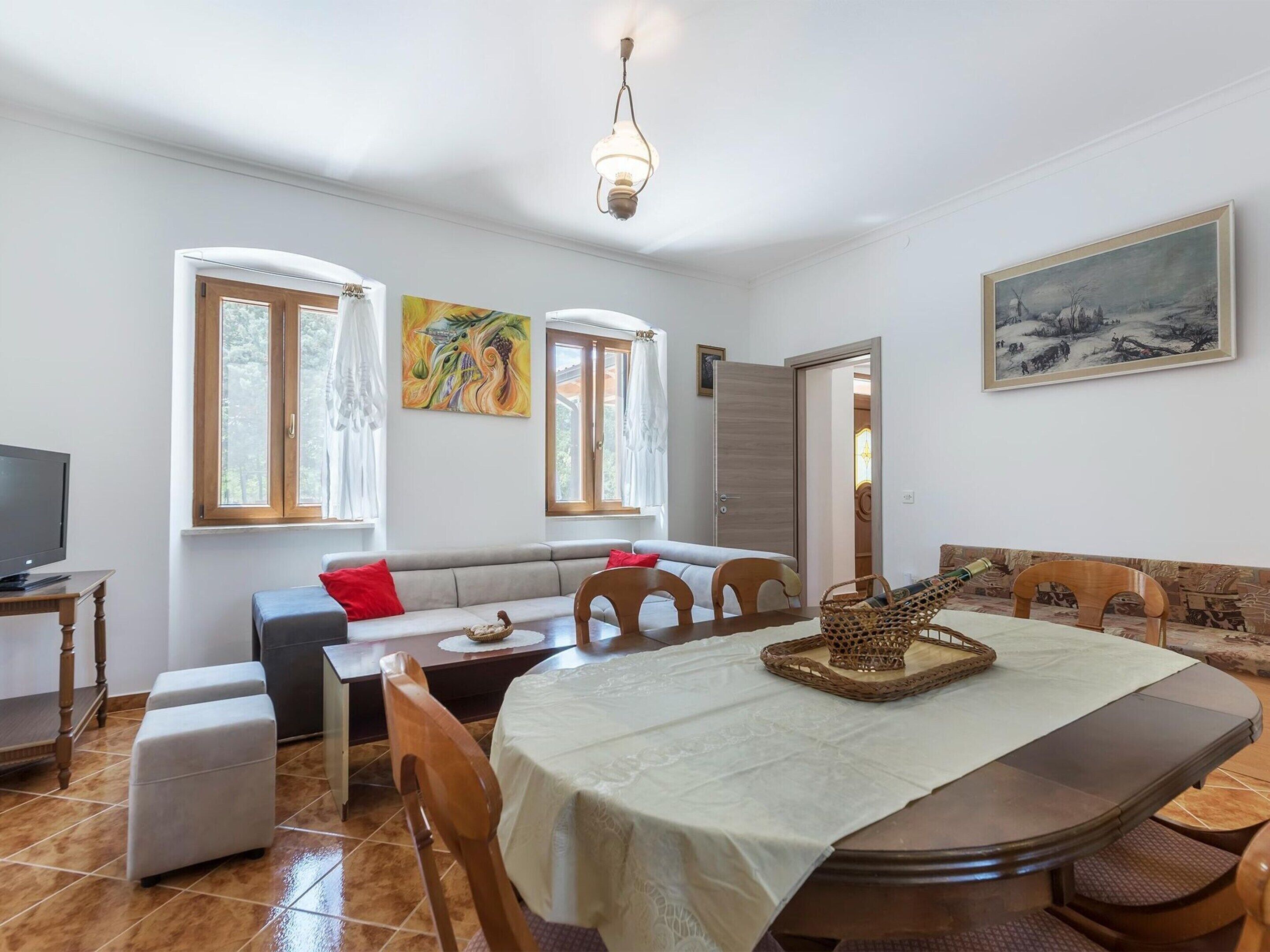 A cozy living room with a sofa, dining table, and chairs. There is a television on a stand, a painting on the wall, and a door leading to another room.