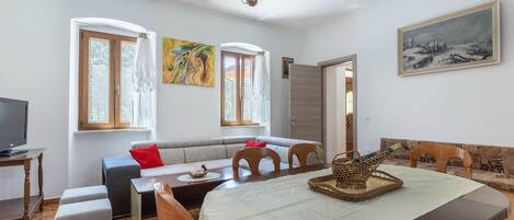 A cozy living room with a sofa, dining table, and chairs. There is a television on a stand, a painting on the wall, and a door leading to another room.