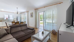 A modern living room with a sofa, a coffee table, and a TV. There is a kitchen area with a window and a dining table. - Large Holiday Home With Swimming Pool (Labin)