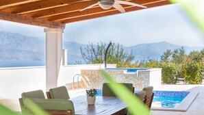 Eine Terrasse im Freien mit Holztisch und -stühlen, einem Pool und einer Bergkulisse.