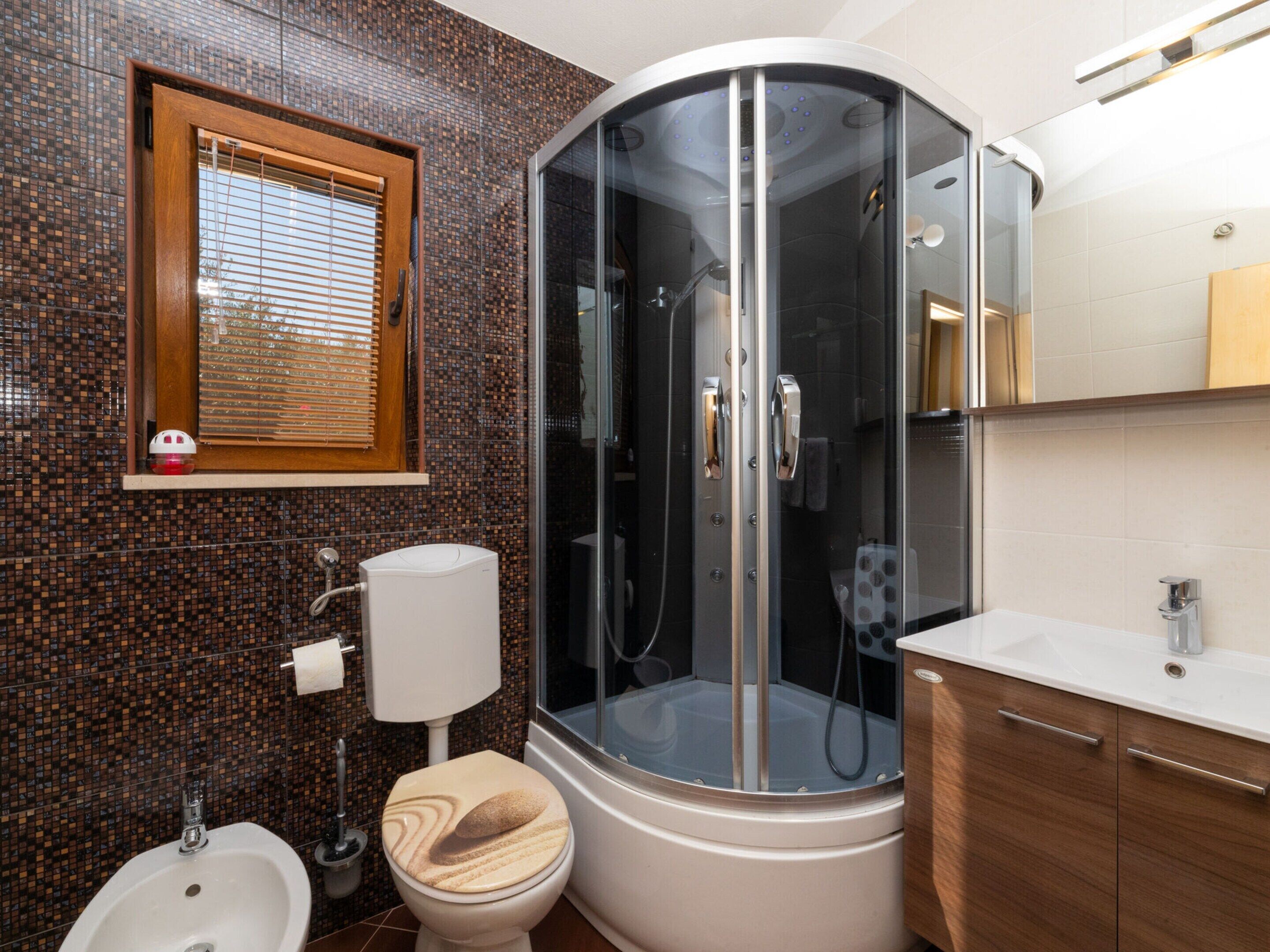 A modern bathroom with a glass-enclosed shower, a toilet with a wooden seat, a bidet, and a sink with a mirror.