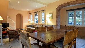 Ein Essbereich mit Holztisch und Korbstühlen, einer Couch und einem Fernseher in einem Raum mit Treppe und großen Fenstern.