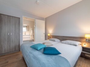 Quarto de hotel moderno com uma cama grande, duas mesas de cabeceira com luminárias, um guarda-roupa e um banheiro visível através de uma porta aberta.