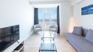 A modern living room with a large TV, a glass coffee table, two armchairs, a sofa, and a view of the beach through large windows. - Modern Apartment on the Seafront (Middelkerke)