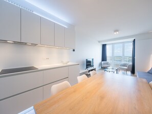 A modern kitchen with white cabinets, a wooden dining table, and a TV in the living area with a balcony view. - Modern Apartment on the Seafront (Middelkerke)