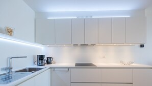 A modern kitchen with white cabinets, a stainless steel sink, a coffee maker, and a wooden dining table. - Modern Apartment on the Seafront (Middelkerke)