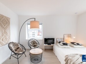 Ein modernes Schlafzimmer mit Bett, zwei Stühlen, einem kleinen Tisch, einem Fernseher und einem Fenster mit Blick auf die Stadt.