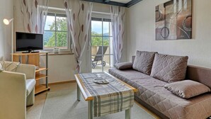 A cozy living room with a sofa, a TV on a stand, a table with a tray, and large windows with curtains. - Holiday Apartment Haus Leutner Bavarian Forest (Bodenmais)