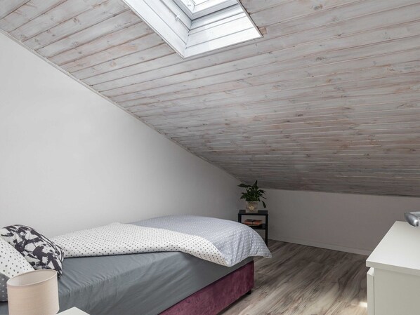 Une chambre avec un plafond en pente, un lit avec une literie grise, une petite table de chevet avec une plante et une commode blanche.