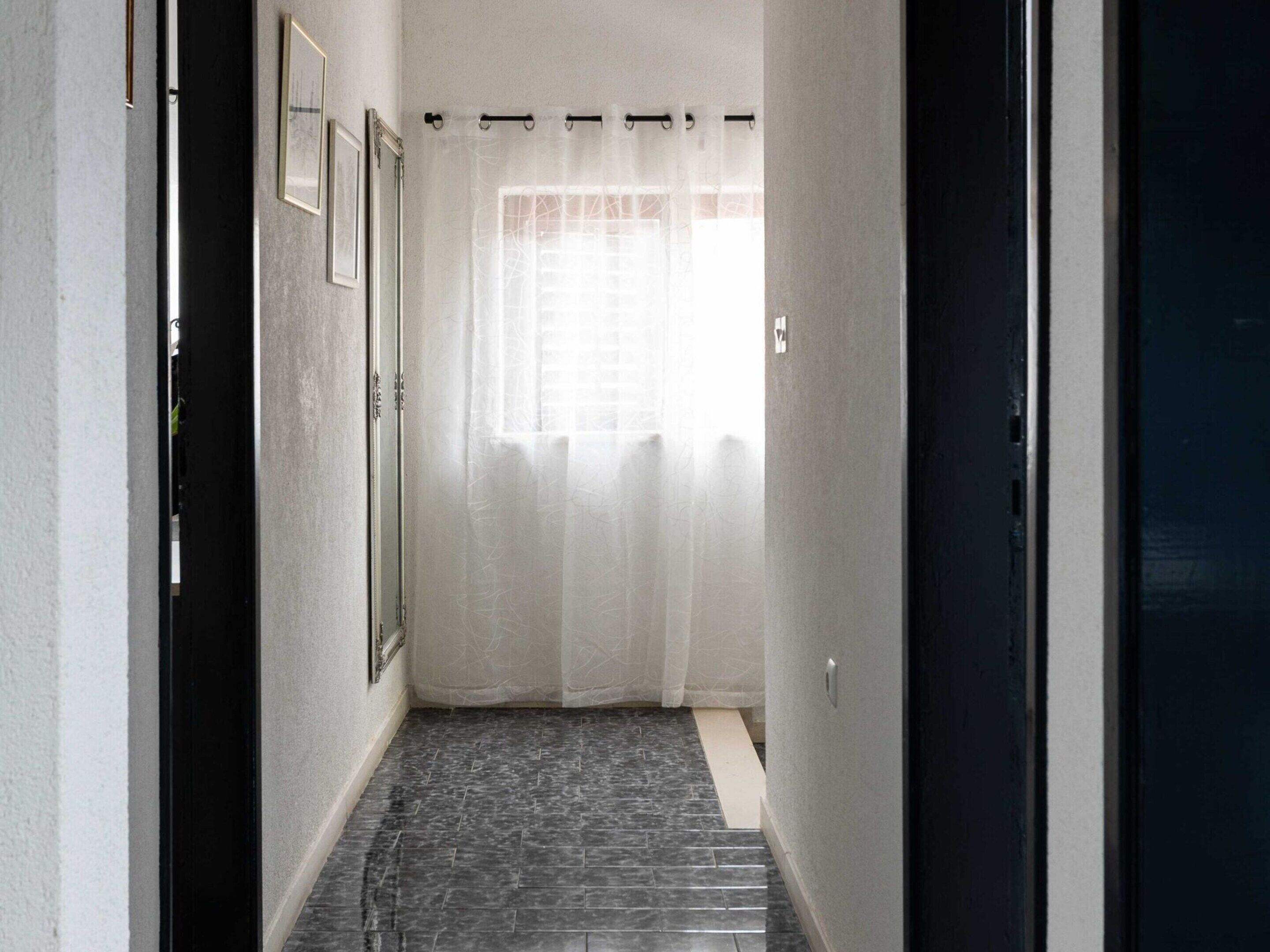 Un couloir doté d’un sol carrelé, une porte noire sur la droite et une fenêtre ornée de rideaux légers au fond.