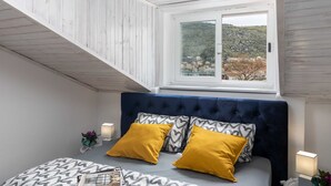 A bedroom with a sloped ceiling, a bed with a dark headboard, two mustard pillows, patterned pillows, a bedside table with a lamp, and a window with a view of a hillside. - Apartment Tete Mare (Dubrovnik)