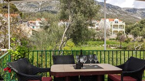 A covered outdoor patio with a table set for four, surrounded by wicker chairs with red cushions, overlooking a mountainous landscape with houses. - Apartment Tete Mare (Dubrovnik)