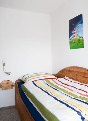 A single bed with a striped bedspread, a wooden headboard, a nightstand, a lamp, and a wall-mounted painting of a lighthouse scene. - Holiday Home Kleiner Friese Dangast (Varel)