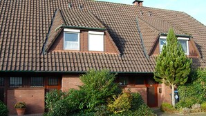 Une maison à deux étages, avec un toit en pente, une façade en briques et un petit jardin à l’avant, agrémenté d’un arbre et de quelques arbustes.