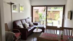 Un salon chaleureux avec un canapé en cuir marron, une petite table ronde et une table à manger avec des chaises. Une grande fenêtre offre une vue sur un jardin et un bâtiment à l’extérieur.