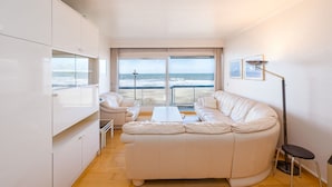 A modern living room with a large white sofa, a coffee table, and a dining table. There is a balcony with a view of the ocean. - Seaview Apartment, Middelkerke (Middelkerke)