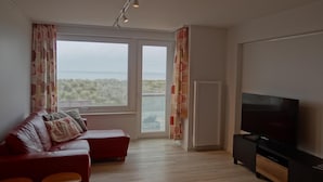 A living room with a red sofa, two wooden stools, a flat-screen TV, and a large window with curtains.