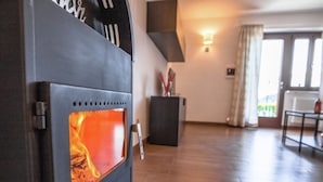 A wood stove with a visible fire, a dark cabinet, a table, and a view of a window with curtains. - Belvilla by OYO Modern Holiday Home With Garden (Ledro)