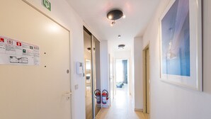 Un pasillo de hotel con paredes blancas, un extintor, letreros de salida de emergencia y un cuadro enmarcado a la derecha.