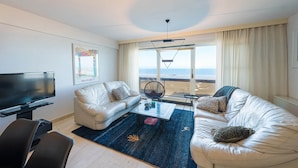 Una sala de estar moderna con un televisor de pantalla plana grande, dos sofás de cuero blanco, una mesa de centro y vistas a la playa a través de amplios ventanales.