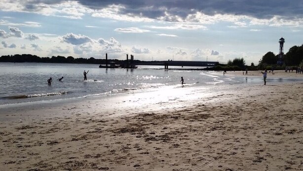 Ein Strand mit einem Fluss im Hintergrund, einer Brücke und einem Leuchtturm.