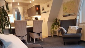 Ein gemütliches Wohnzimmer mit einer grauen Couch, gestreiften Kissen und einem dunklen Sessel. Es gibt einen Essbereich mit zwei gestreiften Stühlen und einem kleinen Tisch. Eine Topfpflanze sorgt für etwas Grün.