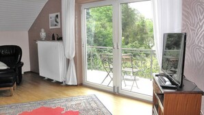 Et værelse med en stor skydedør, der fører ud til en balkon med udsigt over grønne områder. Der er en sort læderstol, en radiator og et fjernsyn på et træskab.