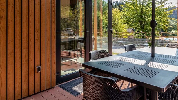 A modern outdoor dining area with a glass door leading to a patio, surrounded by wooden panels and a view of greenery.