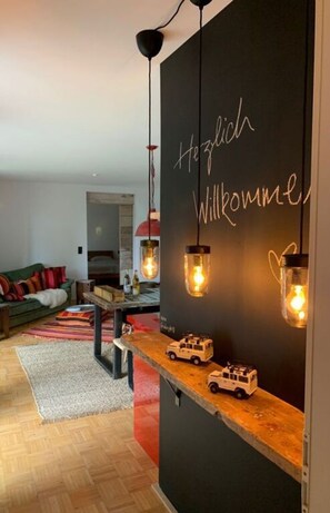 A modern living room with a black wall displaying "Heizlich Willkommen" and a wooden shelf with toy cars. - Ferienwohnung 2 mit Sauna Ferienhaus Poppenhausen (Poppenhausen)